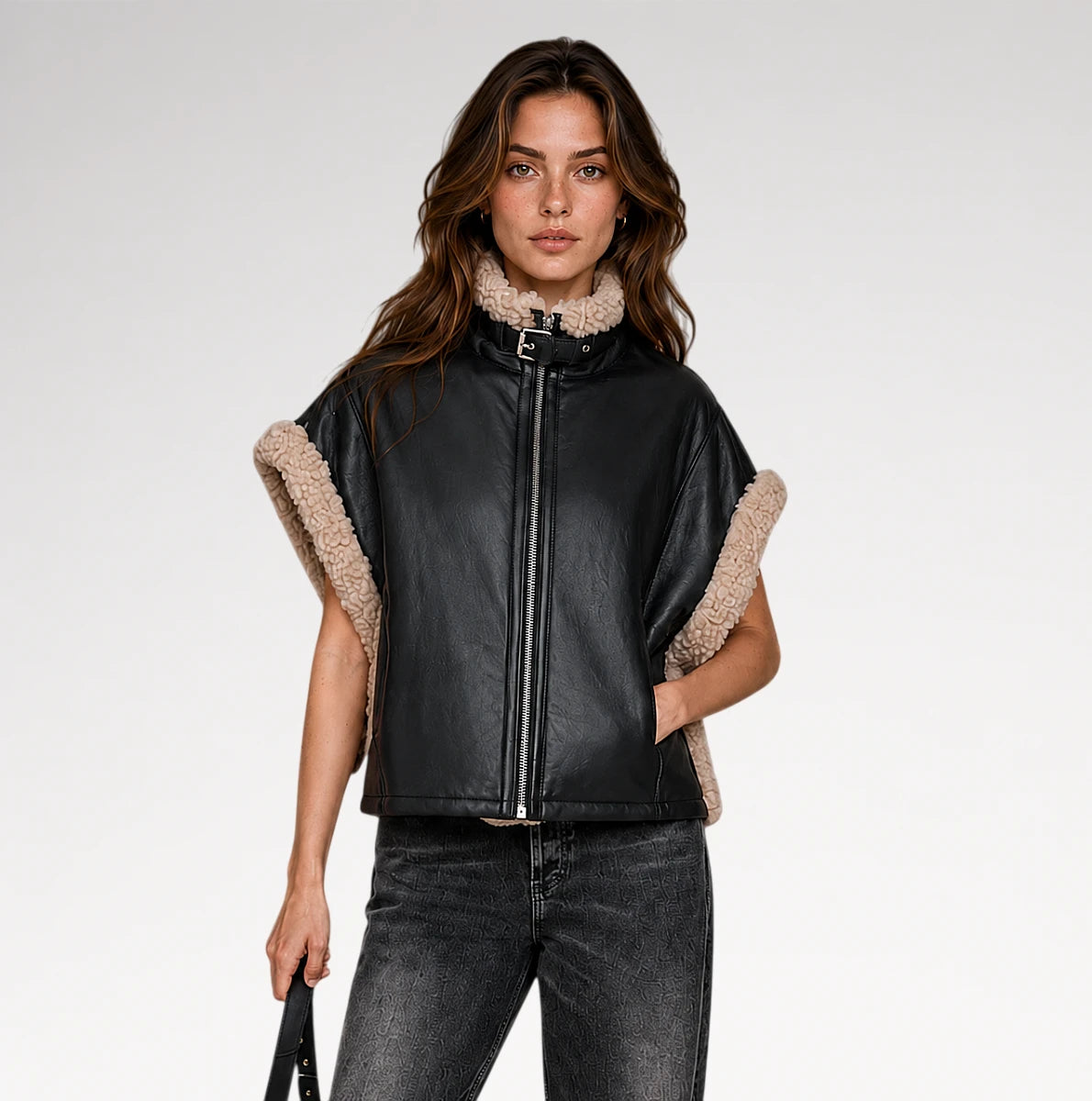 A woman modeling a black faux-leather jacket with beige sherpa lining on the collar and sleeve cuffs, paired with dark gray jeans, against a light gray studio background.