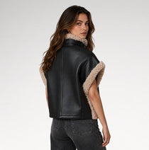 A woman shown from the back wearing a black faux-leather jacket with beige sherpa lining on the collar and sleeve cuffs, paired with dark gray jeans, against a light gray studio background.