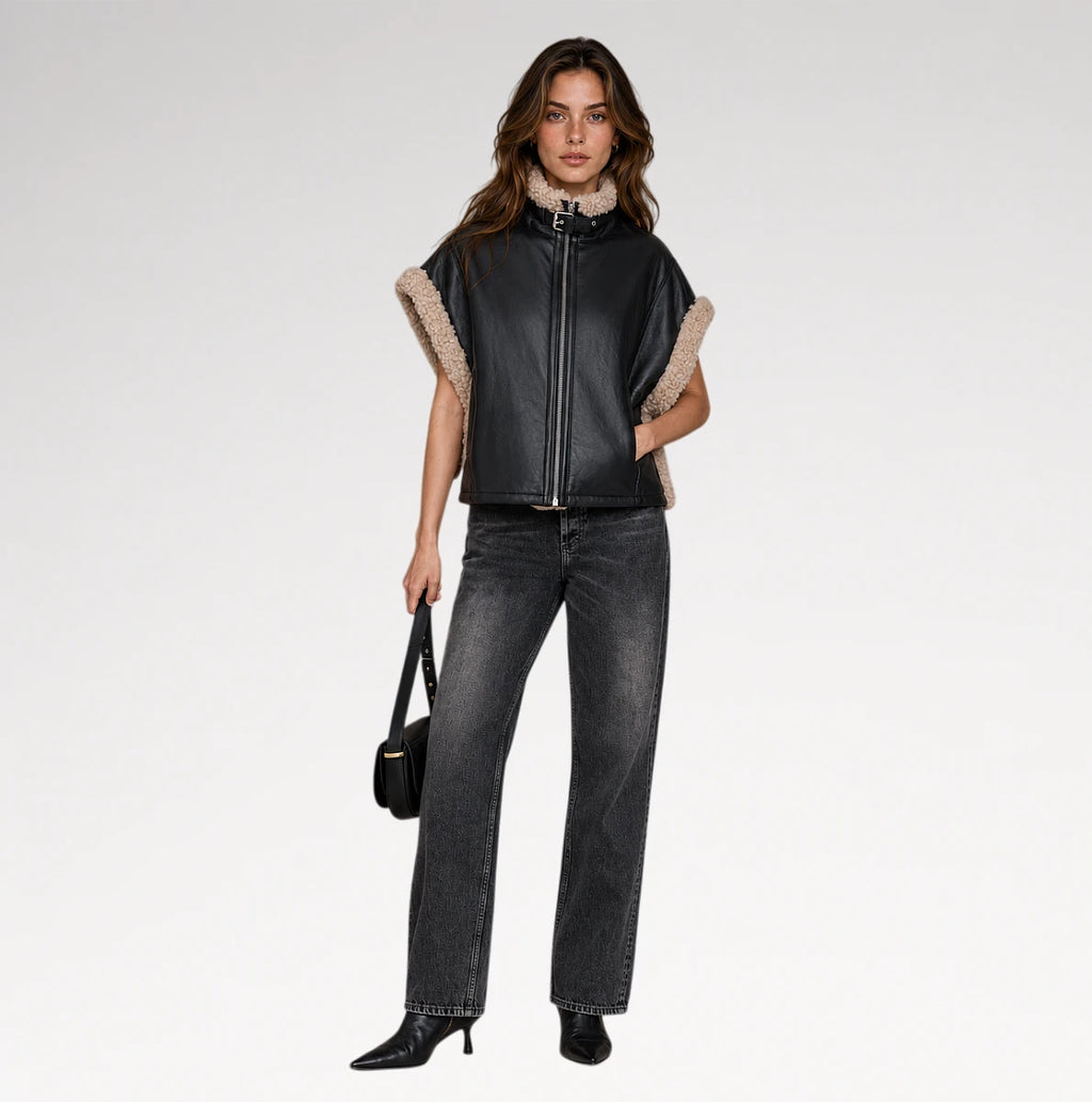 A model stands against a light gray studio background wearing a black leather moto jacket with beige shearling trim on the sleeves and collar, paired with dark gray jeans, black boots, and holding a small black handbag.