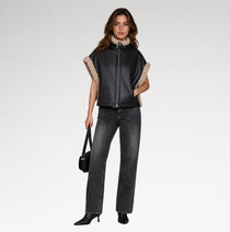 A model stands against a light gray studio background wearing a black leather moto jacket with beige shearling trim on the sleeves and collar, paired with dark gray jeans, black boots, and holding a small black handbag.
