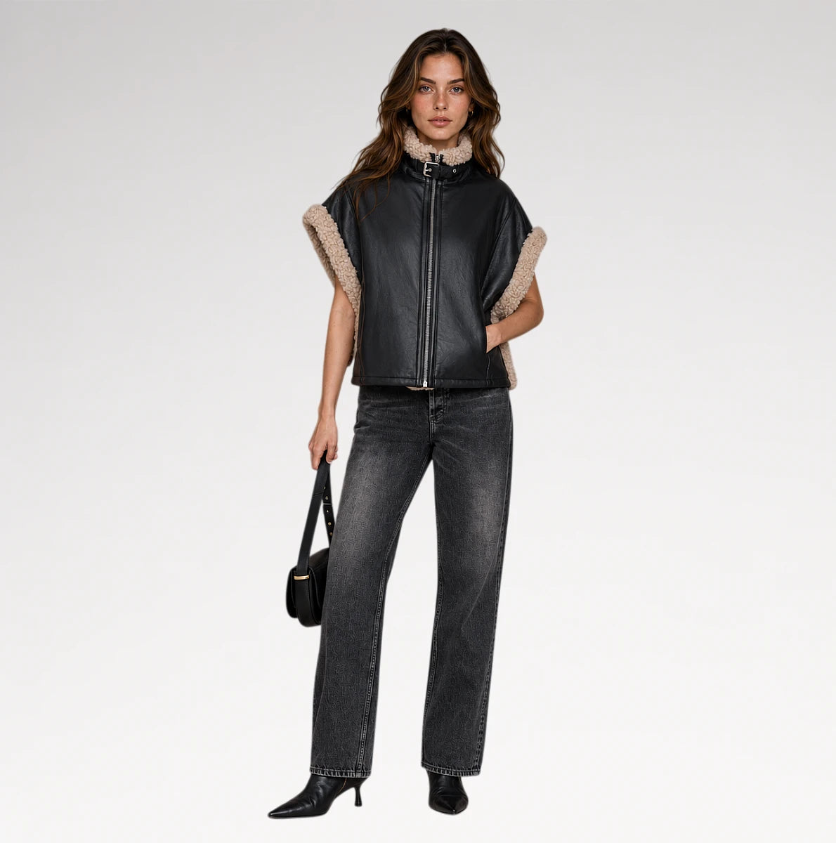 A model stands against a light gray studio background wearing a black leather moto jacket with beige shearling trim on the sleeves and collar, paired with dark gray jeans, black boots, and holding a small black handbag.