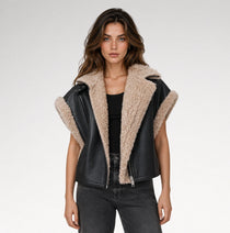 A woman modeling a black faux-leather moto jacket with a beige sherpa fleece lining, worn over a black top and dark gray jeans against a light gray studio background.