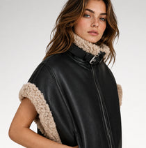 A woman wearing a black faux-leather vest with beige bouclé trim and a matching beige knit neck warmer, photographed against a clean light gray studio background.