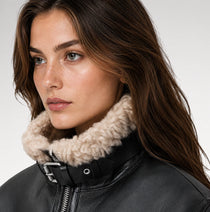 Close-up of a woman wearing a black leather jacket with a thick beige sherpa fleece collar, set against a plain light gray background.
