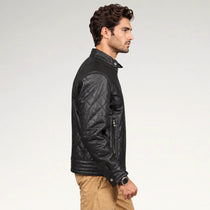 A man in profile wearing a black quilted leather jacket with zippered pockets, paired with tan pants, against a plain light gray studio background.