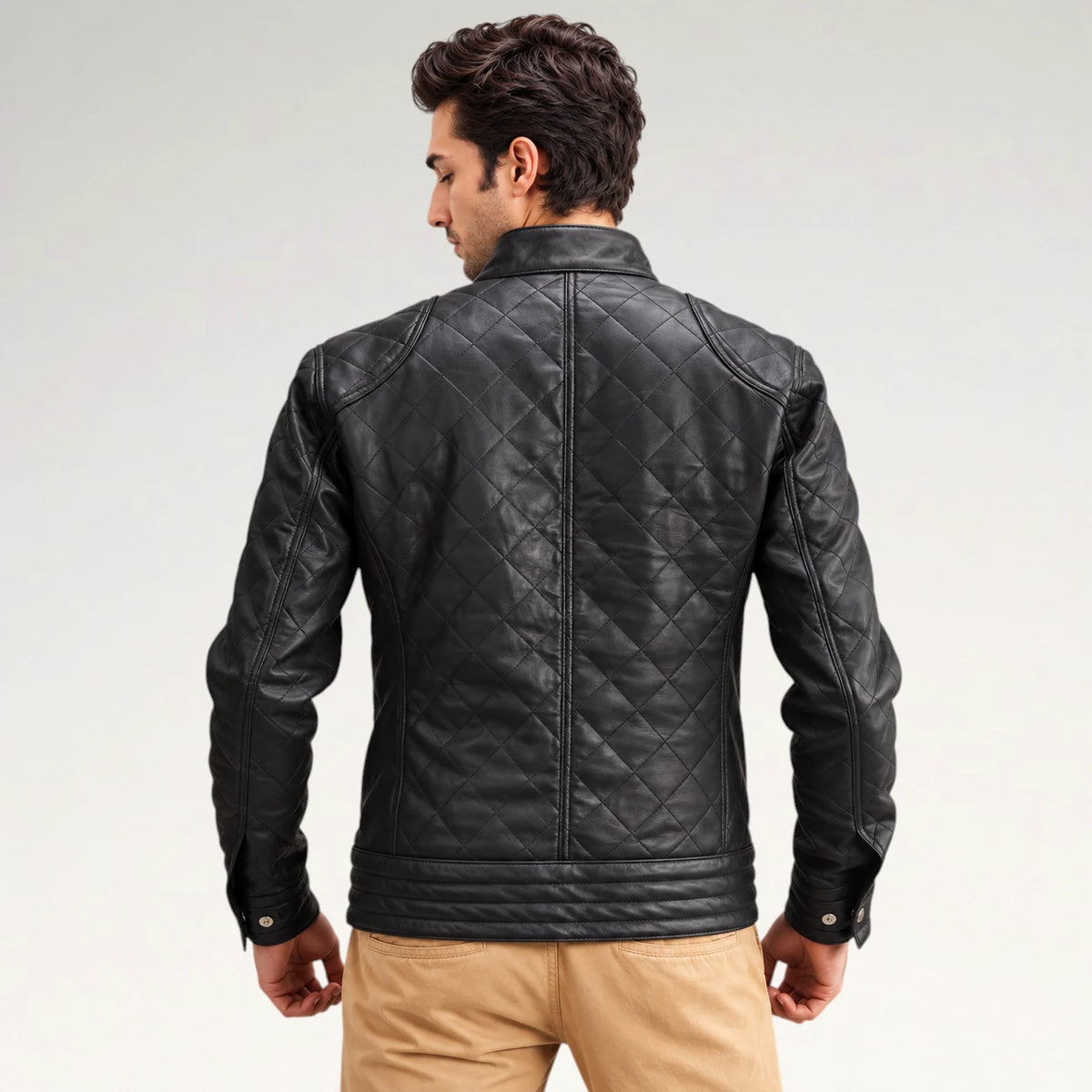 A man viewed from the back wearing a black quilted leather jacket and tan pants, photographed against a clean light gray studio background.