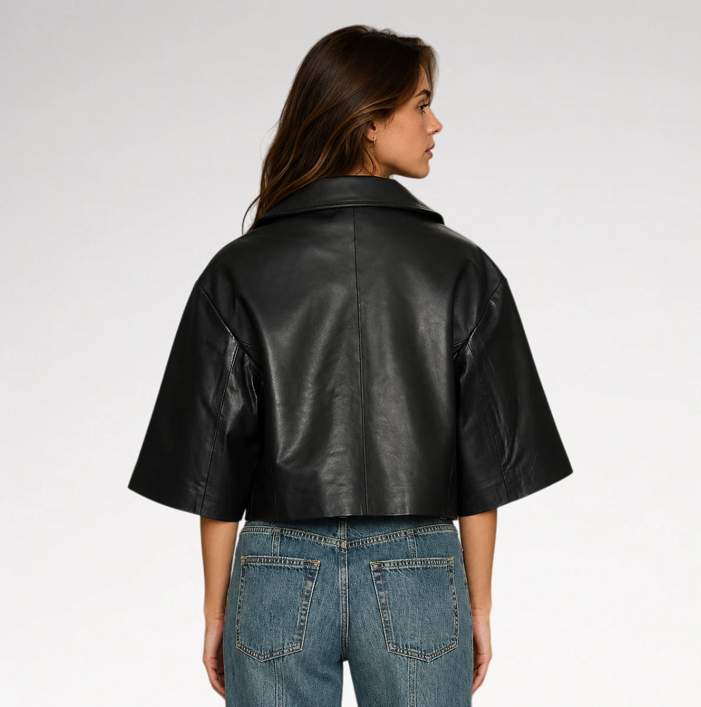 A woman shown from the back wearing a cropped black leather jacket with wide short sleeves and blue jeans, photographed against a light gray studio background.