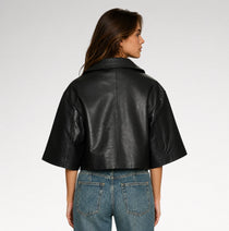 A woman shown from the back wearing a cropped black leather jacket with wide short sleeves and blue jeans, photographed against a light gray studio background.