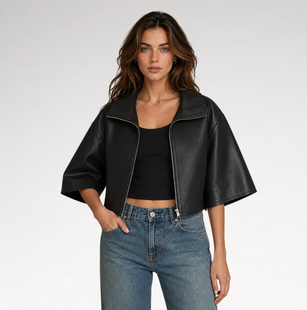 A woman modeling a black cropped faux-leather jacket with wide three-quarter sleeves, worn over a black crop top and blue jeans, against a light gray studio background.