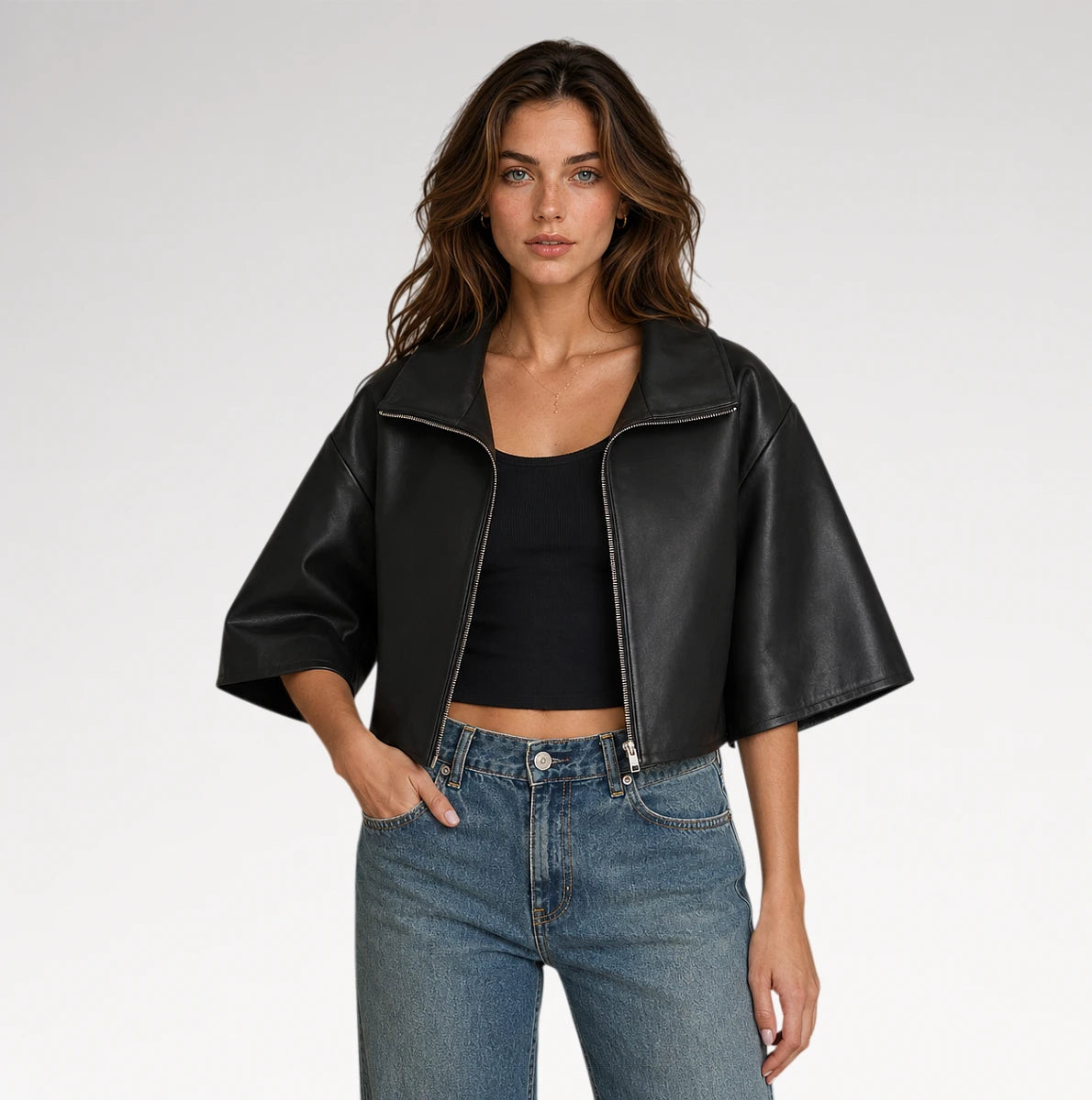 A woman modeling a black cropped faux-leather jacket with wide three-quarter sleeves, worn over a black crop top and blue jeans, against a light gray studio background.