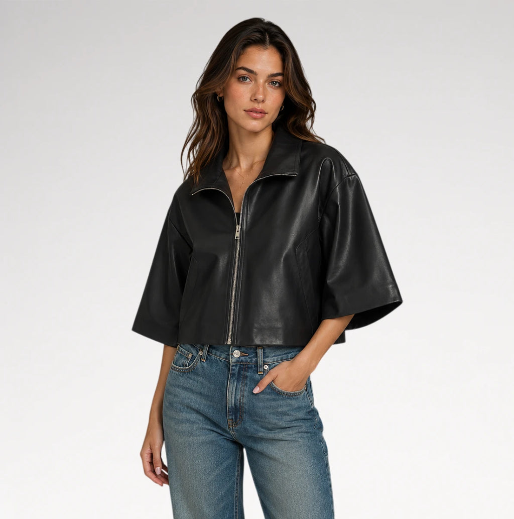 A woman modeling a black cropped faux-leather jacket with wide short sleeves and a front zipper, paired with blue jeans, against a light gray studio background.