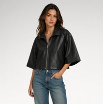 A woman modeling a black cropped faux-leather jacket with wide short sleeves and a front zipper, paired with blue jeans, against a light gray studio background.
