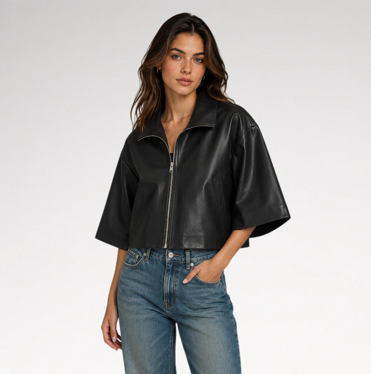 A woman modeling a black cropped faux-leather jacket with wide short sleeves and a front zipper, paired with blue jeans, against a light gray studio background.