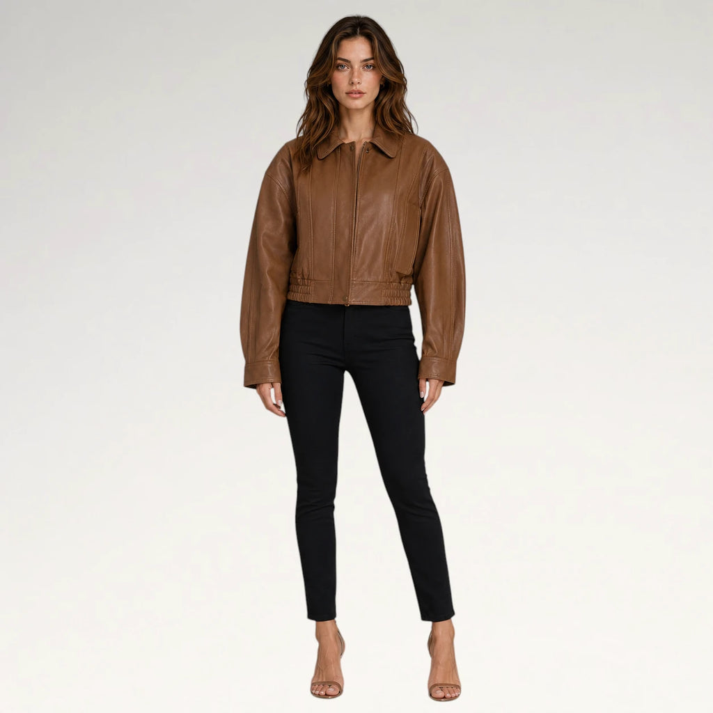 A woman modeling a cropped brown leather jacket with black skinny pants and nude high-heeled sandals, standing against a light gray studio background.