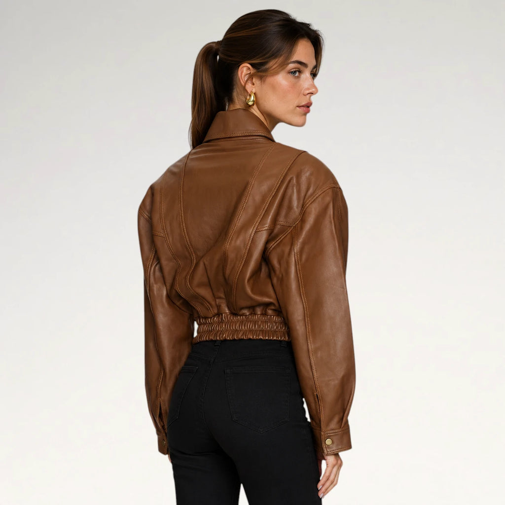 A woman shown from the back wearing a cropped brown leather jacket with a gathered elastic waist and black pants, photographed against a clean light gray studio background.
