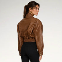A woman shown from the back wearing a cropped brown leather jacket with a gathered elastic waist and black pants, photographed against a clean light gray studio background.