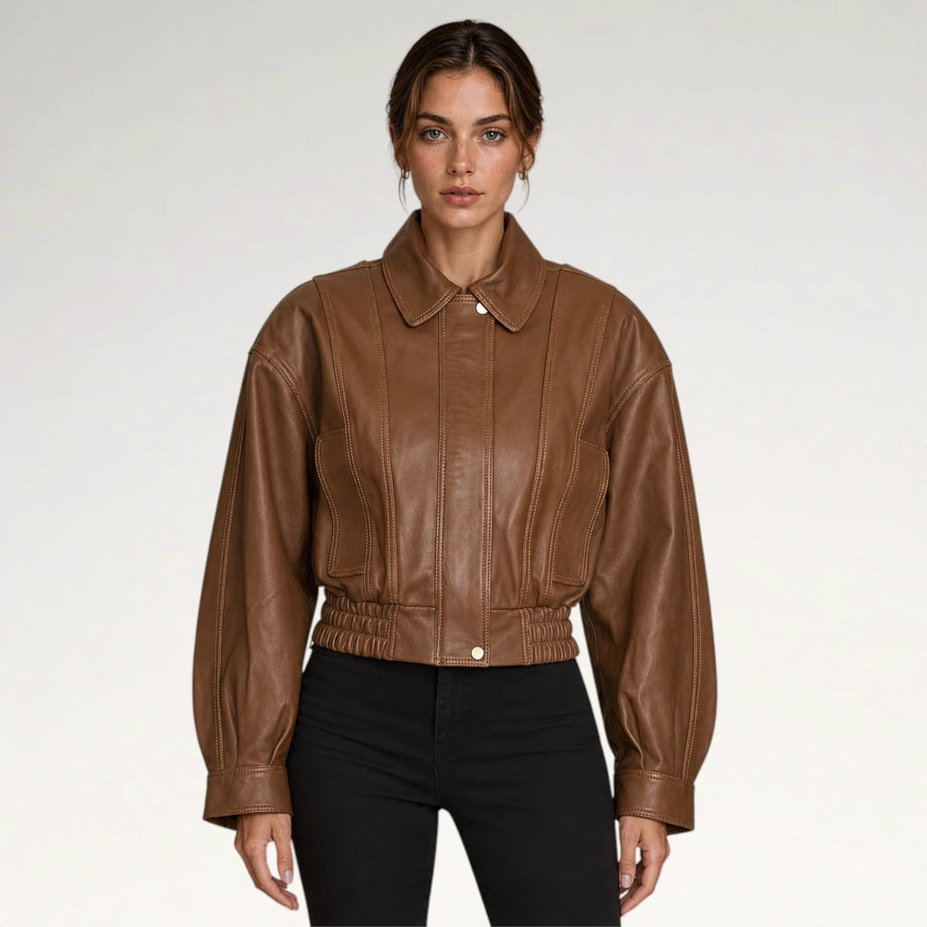 A woman modeling a cropped brown leather jacket with a pointed collar, front button closure, and gathered elastic waist, paired with black pants against a light gray studio background.