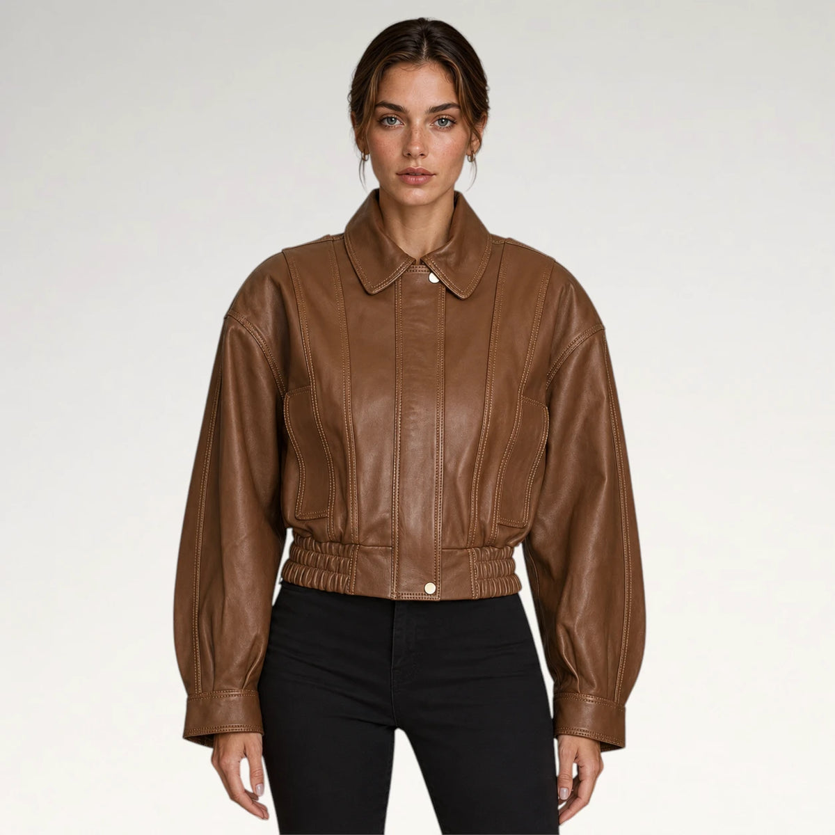 A woman modeling a cropped brown leather jacket with a pointed collar, front button closure, and gathered elastic waist, paired with black pants against a light gray studio background. Main image