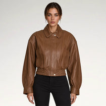 A woman modeling a cropped brown leather jacket with a pointed collar, front button closure, and gathered elastic waist, paired with black pants against a light gray studio background.