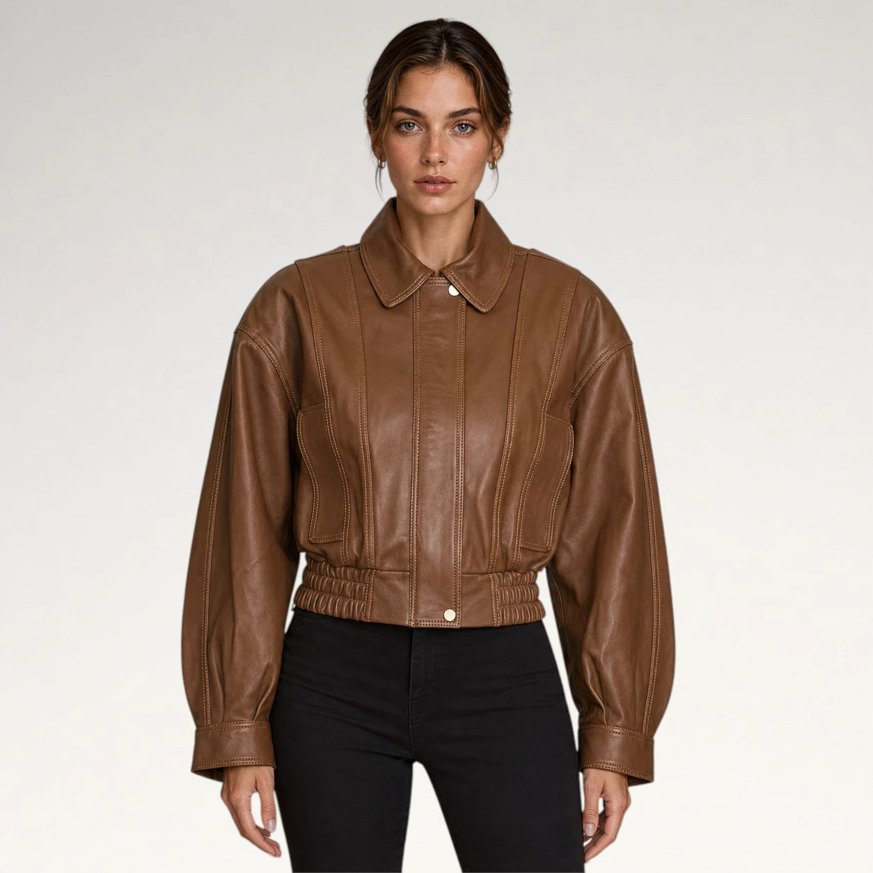 A woman modeling a cropped brown leather jacket with a pointed collar, front button closure, and gathered elastic waist, paired with black pants against a light gray studio background.