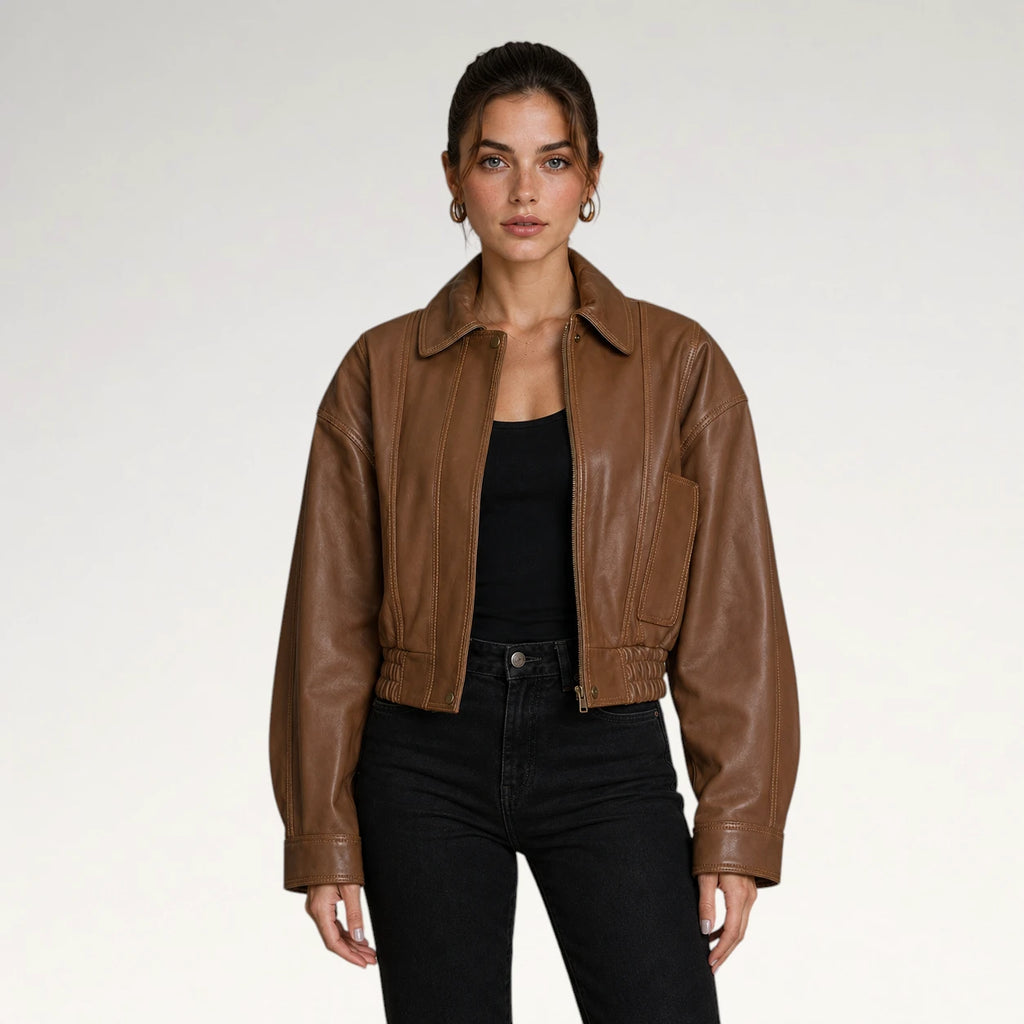 A woman modeling a cropped brown faux-leather jacket over a black top and black jeans, photographed against a clean light gray studio background.
