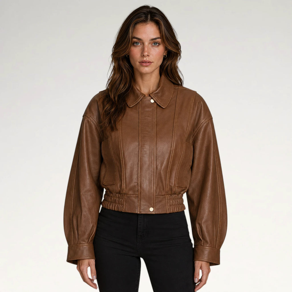 A woman modeling a cropped brown faux-leather jacket with a pointed collar, button front, and gathered elastic hem, paired with black pants against a light gray studio background.