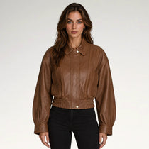 A woman modeling a cropped brown faux-leather jacket with a pointed collar, button front, and gathered elastic hem, paired with black pants against a light gray studio background.