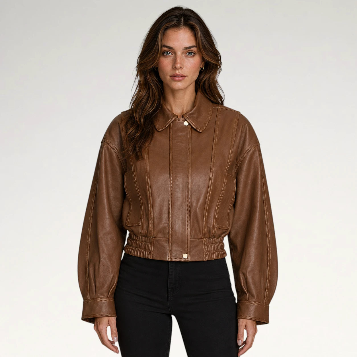 A woman modeling a cropped brown faux-leather jacket with a pointed collar, button front, and gathered elastic hem, paired with black pants against a light gray studio background.