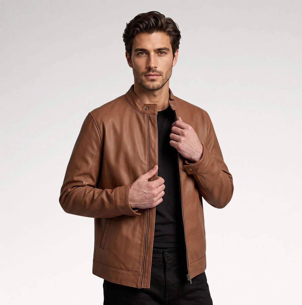 A man wearing a brown leather jacket over a black shirt and black pants, standing against a light gray studio background.