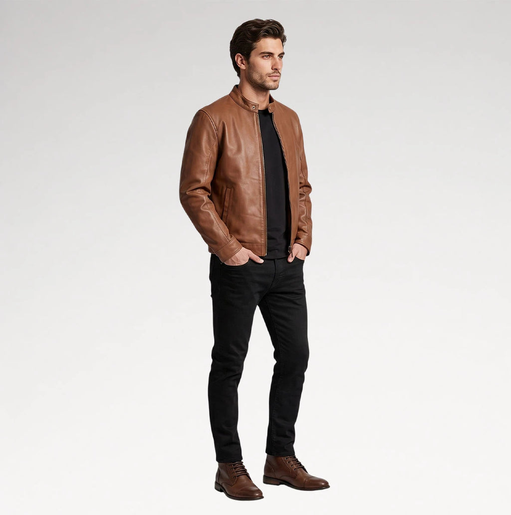 A man wearing a brown leather jacket over a black shirt and black jeans, standing against a light gray studio background.