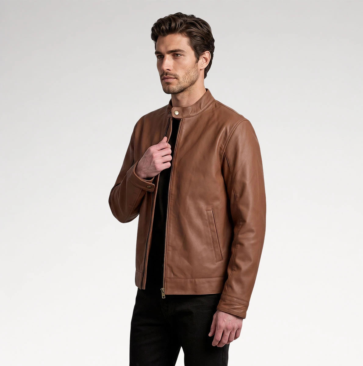 A man wearing a brown leather jacket over a black shirt and black pants, standing against a light gray studio background.