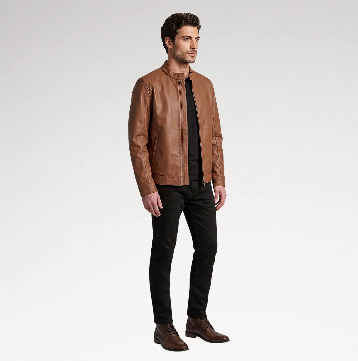 A man modeling a brown leather jacket, black shirt, black pants, and brown boots against a light gray studio background.