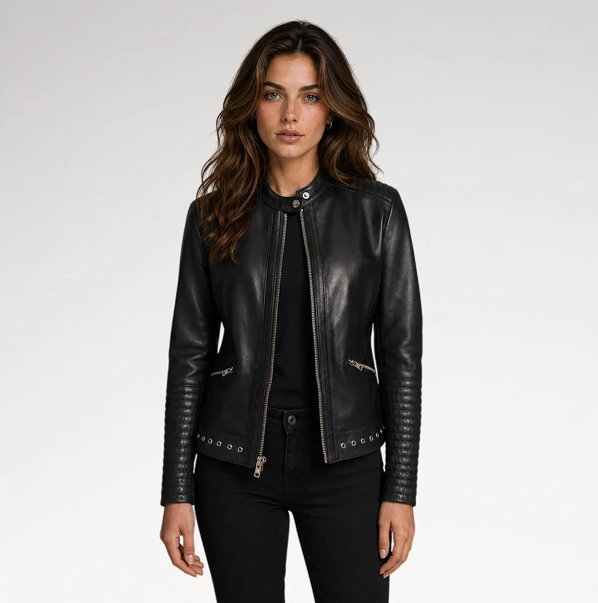 A woman modeling a black leather moto jacket with silver-tone hardware, zippers, and sleeve studs, paired with black pants against a light gray studio background.
