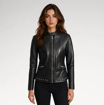 A woman modeling a black faux-leather moto jacket with silver-tone hardware, including a front zipper, zippered hand pockets, and studded detailing along the hem and sleeve cuffs, set against a plain light gray studio background.