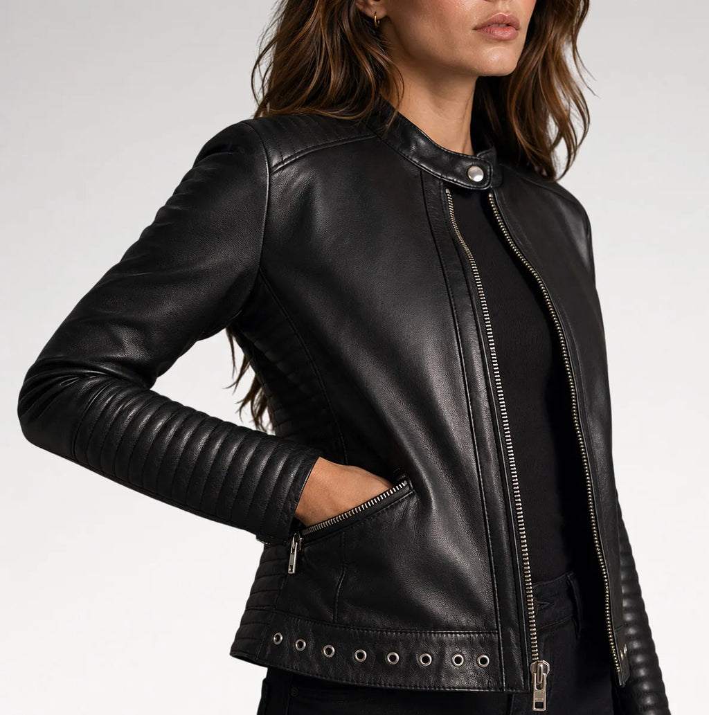 A woman wearing a black leather moto jacket with silver zippers, side pockets, and metal grommets, shown in a close-up studio shot against a light gray background.