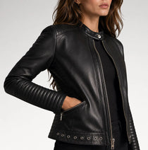 A woman wearing a black leather moto jacket with silver zippers, side pockets, and metal grommets, shown in a close-up studio shot against a light gray background.