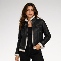 A woman modeling a black faux-leather shearling jacket with a white sherpa lining, worn over a black top and black jeans against a light gray studio background.