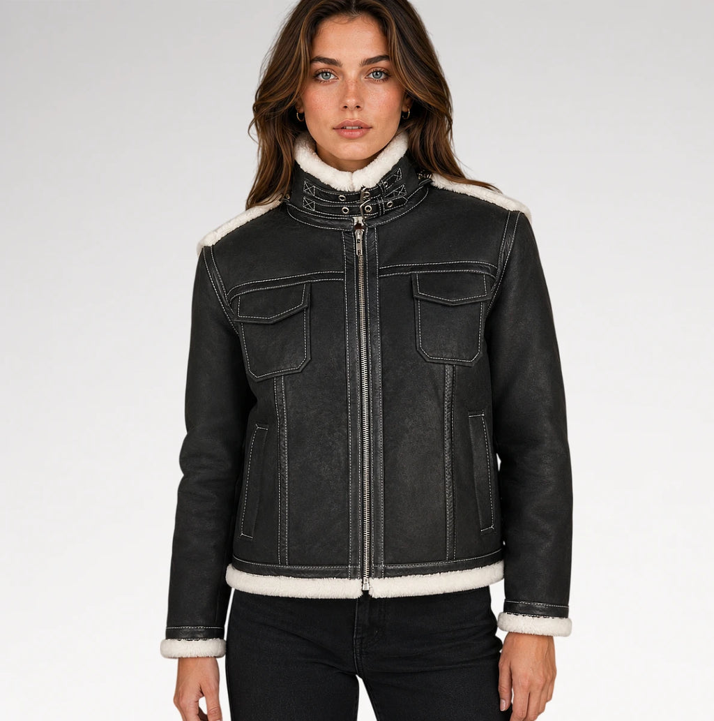 A woman modeling a black faux-leather shearling jacket with a high collar, white sherpa lining, and contrast stitching, paired with black jeans against a light gray studio background.