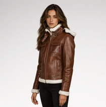 A woman wearing a brown leather aviator-style jacket with white shearling trim on the hood, collar, cuffs, and hem, paired with black pants, photographed against a light gray studio background.