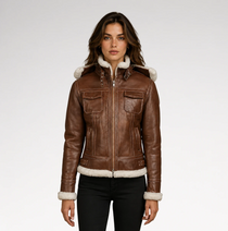 A woman modeling a brown leather aviator-style jacket with white shearling lining on the hood, collar, cuffs, and hem, paired with black pants against a light gray studio background.