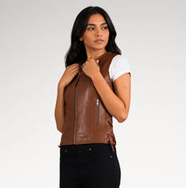 A woman modeling a brown faux leather moto vest with side laces and a front zipper, worn over a white t-shirt and black jeans against a light gray background.