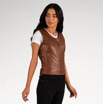 A woman modeling a brown faux leather motorcycle vest with a front zipper, side lacing, and zippered pockets, worn over a white t-shirt and black jeans against a light gray studio background.