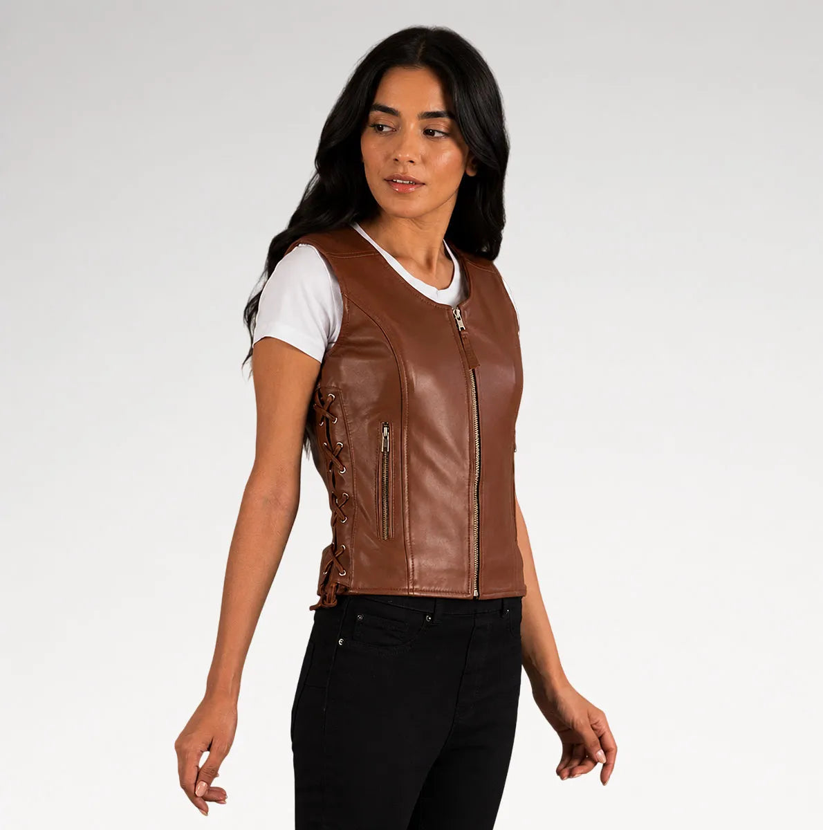 A woman modeling a brown faux leather motorcycle vest with a front zipper, side lacing, and zippered pockets, worn over a white t-shirt and black jeans against a light gray studio background.
