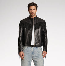 A man modeling a black leather jacket with fringe detailing across the chest, paired with a white t-shirt and light blue jeans, against a plain light gray studio background.