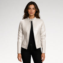 A woman modeling a white faux leather moto jacket with diagonal quilted stitching on the chest and sleeves, paired with a black top and black pants against a light gray studio background.