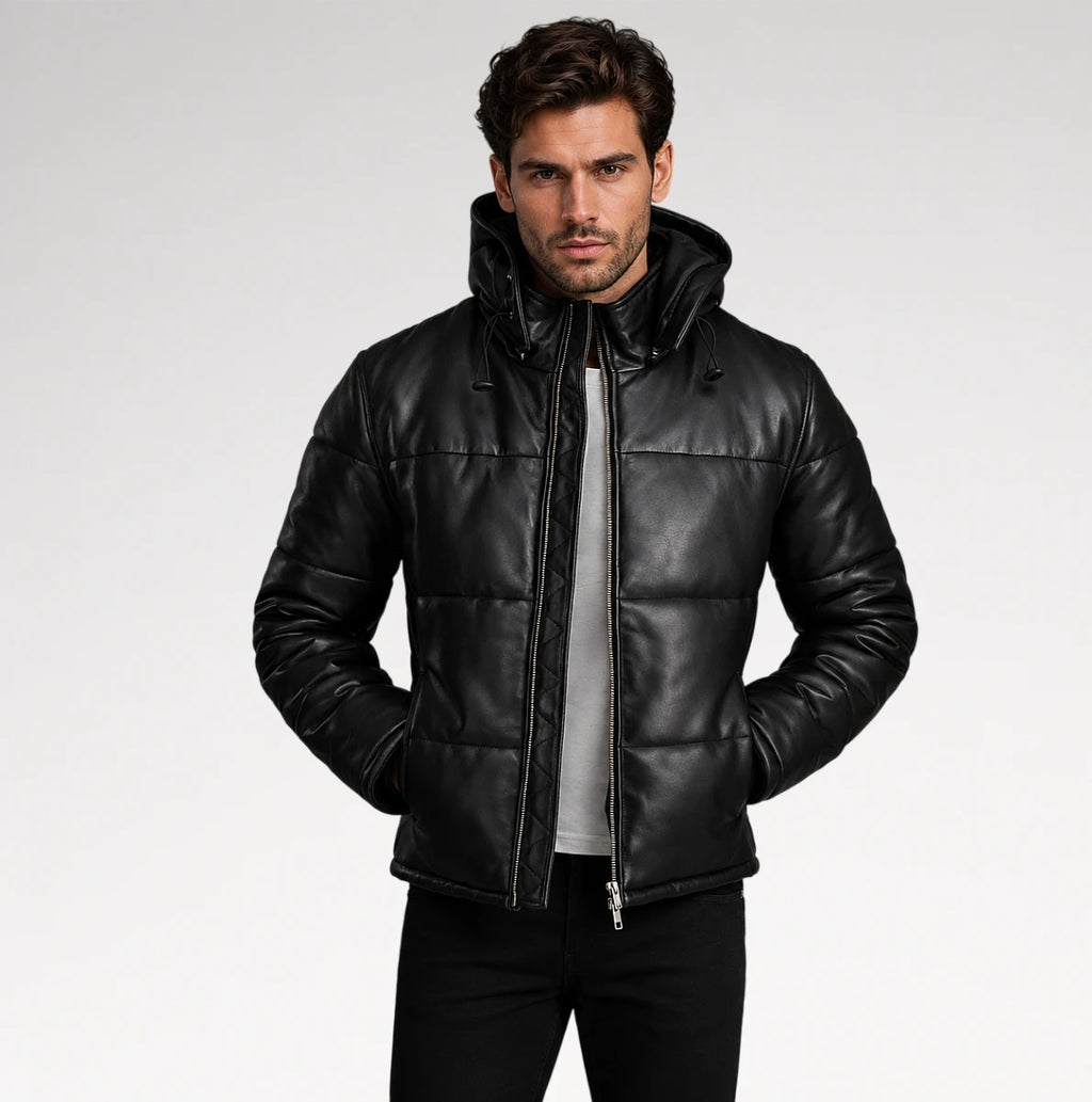 A man wearing a black quilted leather hooded jacket over a gray shirt and black pants, standing against a light gray studio background.