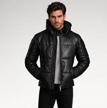 A man wearing a black quilted leather hooded jacket over a gray shirt and black pants, standing against a light gray studio background.