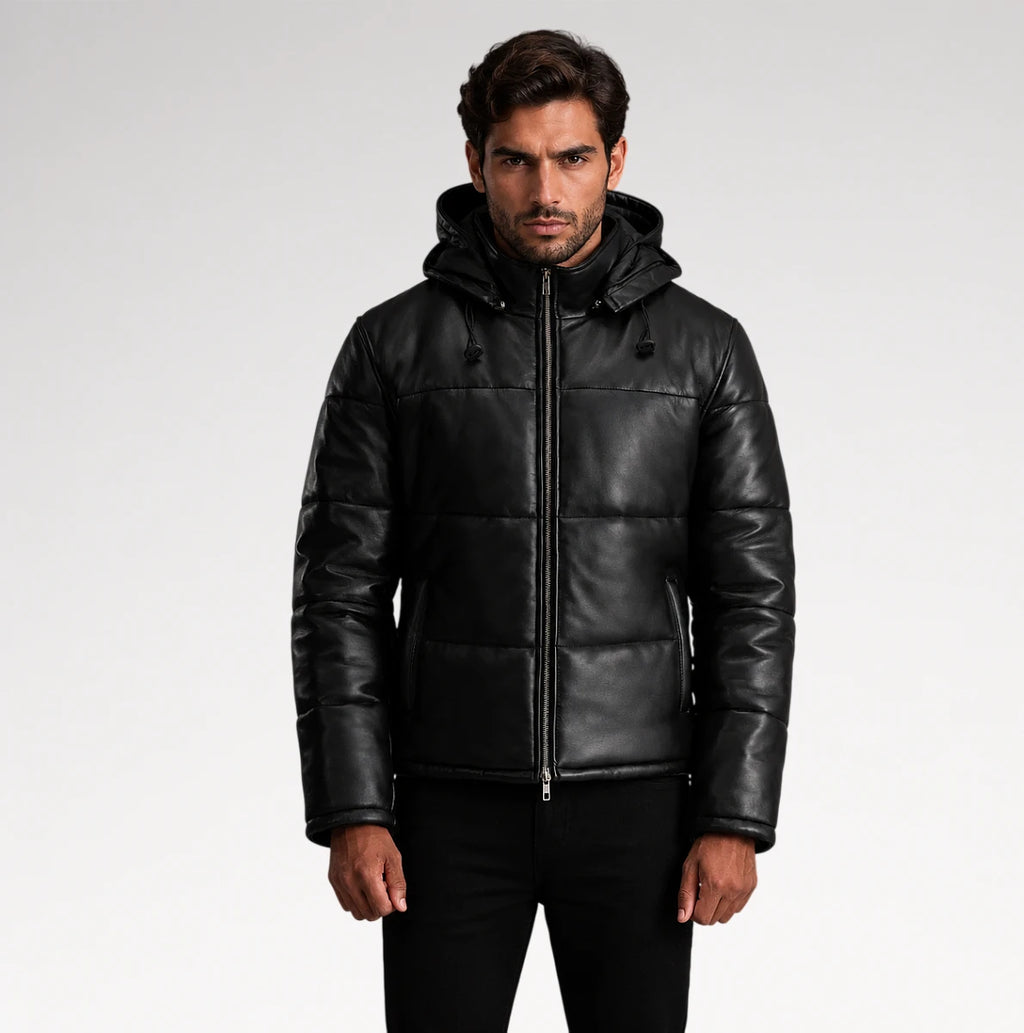 A man wearing a black hooded puffer jacket made of smooth leather or faux leather, paired with black pants, photographed against a light gray studio background.