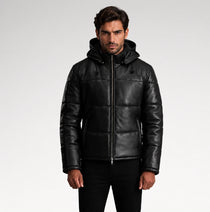 A man wearing a black hooded puffer jacket made of smooth leather or faux leather, paired with black pants, photographed against a light gray studio background.