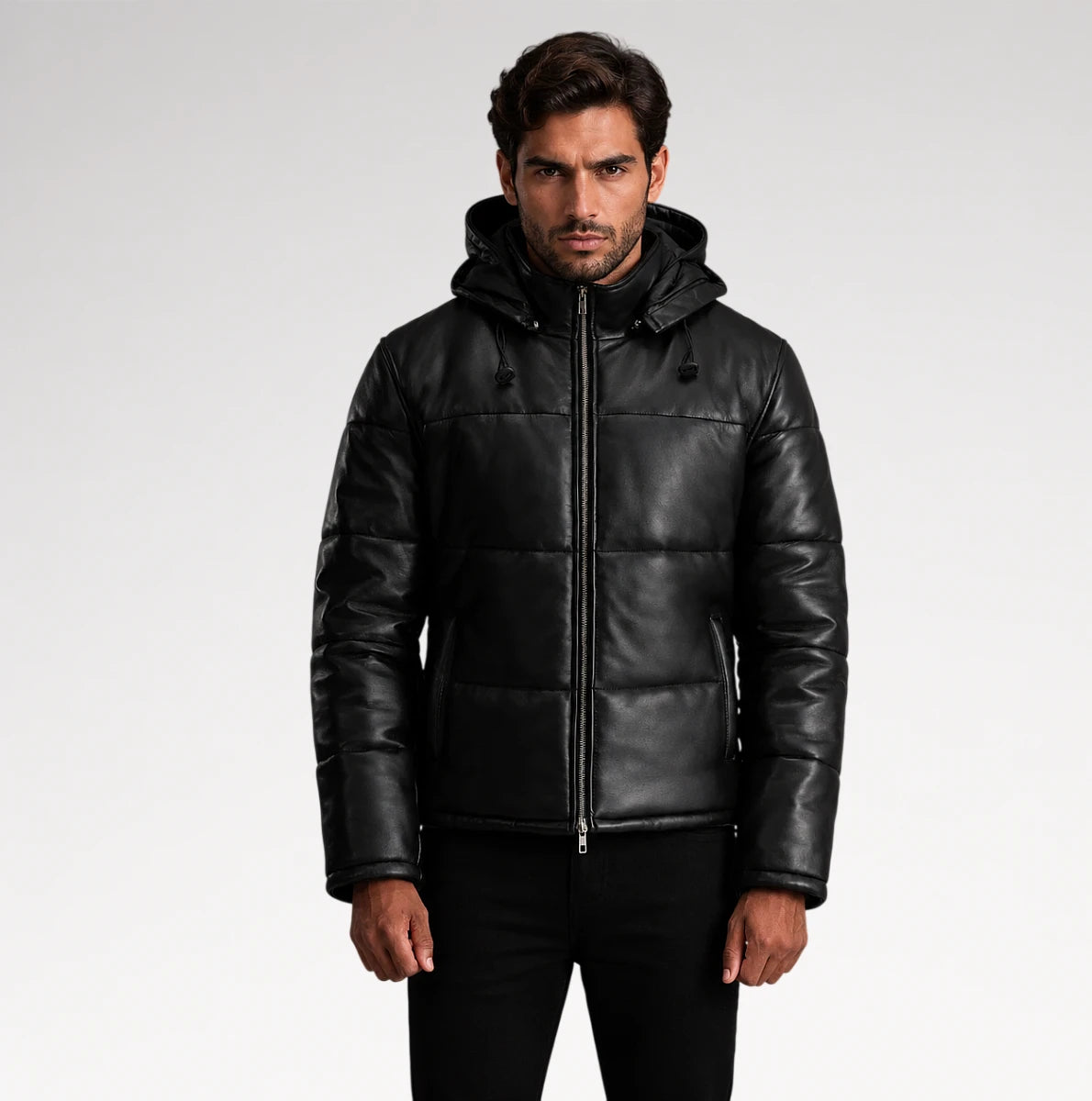 A man wearing a black hooded puffer jacket made of smooth leather or faux leather, paired with black pants, photographed against a light gray studio background.